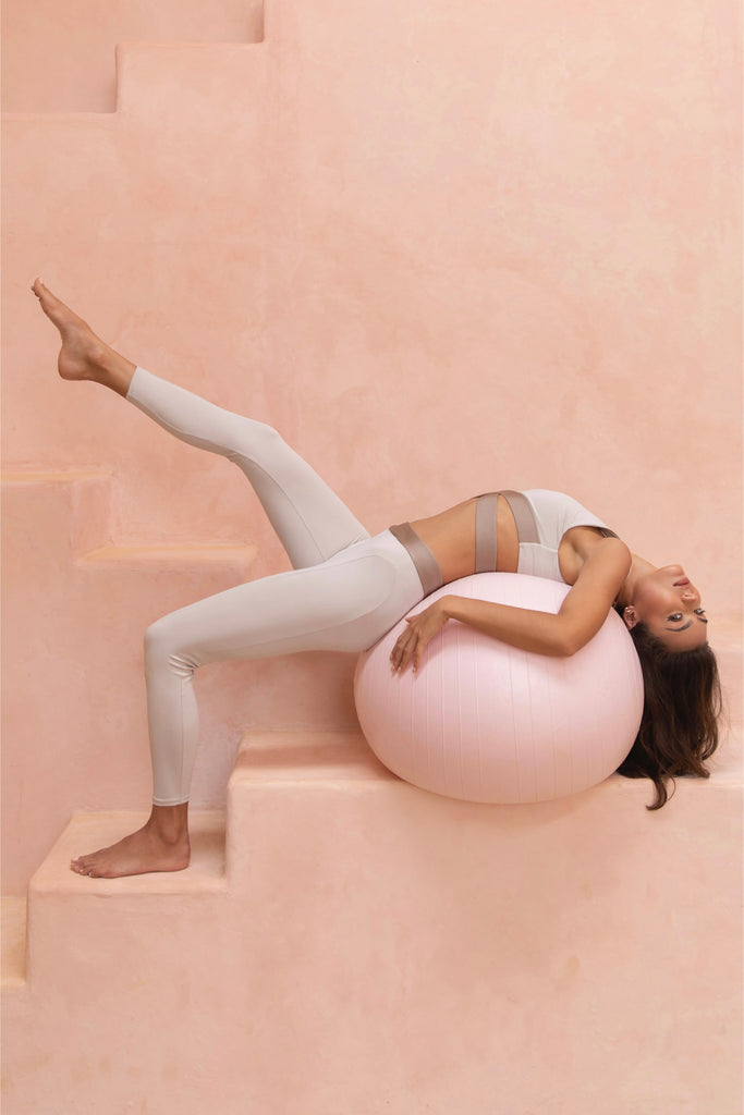 Aesthethic image of a person in the Athena Set (sports bra & leggings) in bone color posing bent over backwards on a pink exercise ball with one leg raised.